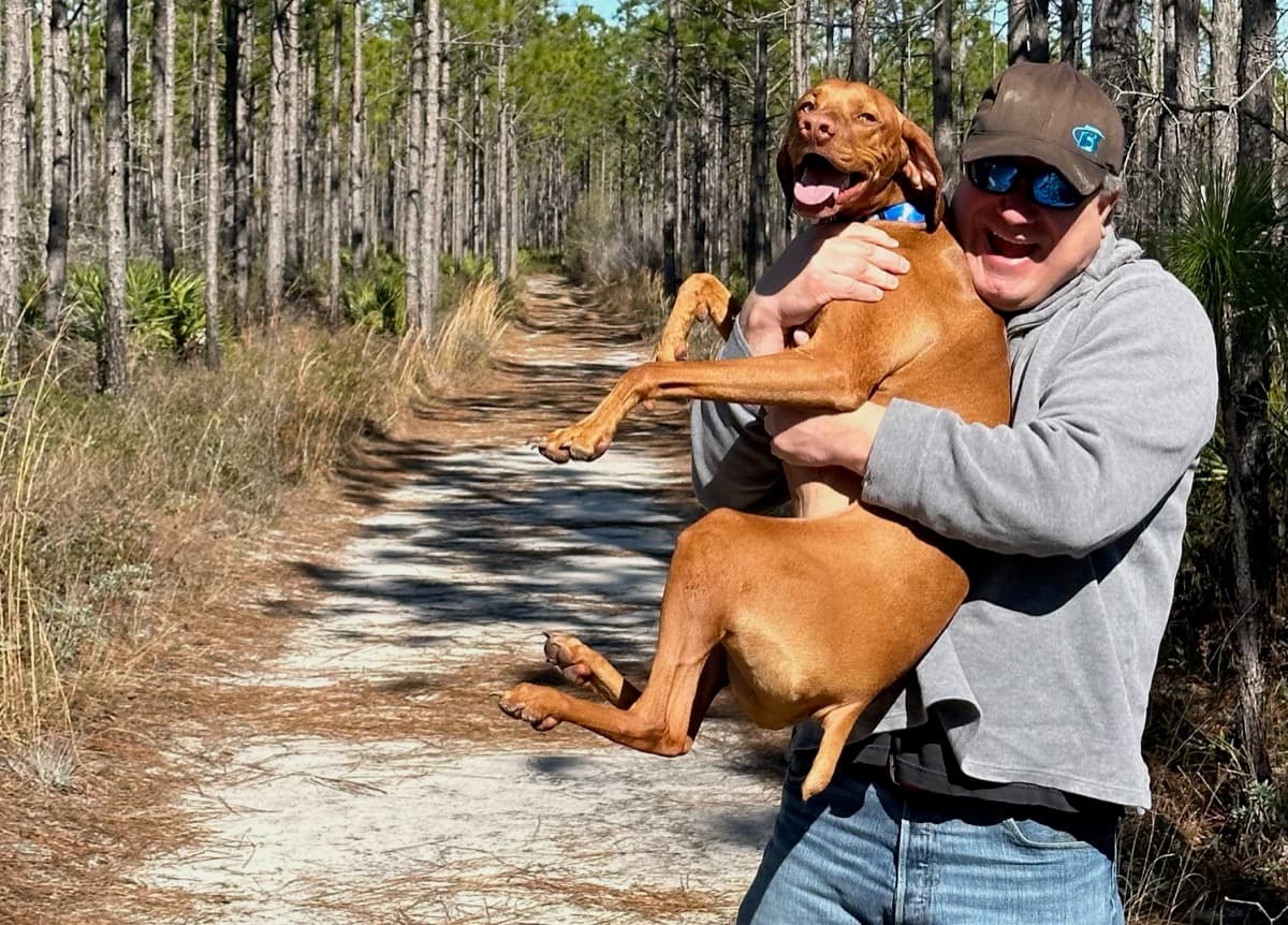 Garett Huber holding Buda, his Vizsla, on a sandy trail through longleaf pines near Santa Rosa Beach, Florida